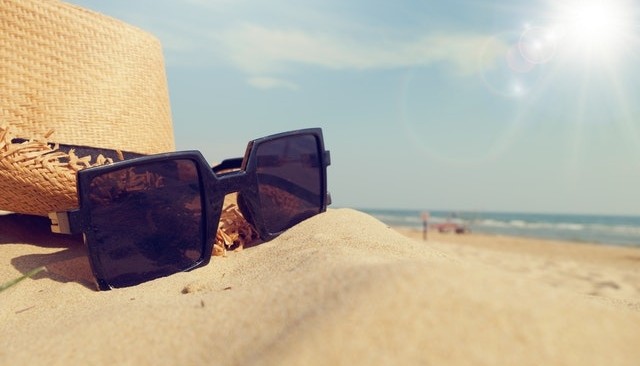 Kolorowa fotografia. Na plaży leży kapelusz i okulary przeciwsłoneczne. W tle słońce, błękitne niebo i morze.