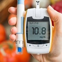 Kolorowa fotografia. Kobieta trzyma w prawej dłoni glukometr. Na drugim planie (w tle) owoce i warzywa.