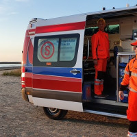 Kolorowa fotografia. Na plaży stoi ambulans z otwartymi drzwiami. W środku ambulansu i przed karetką stoją ratownicy.