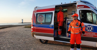 Kolorowa fotografia. Na plaży stoi ambulans z otwartymi drzwiami. W środku ambulansu i przed karetką stoją ratownicy.