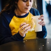 Kolorowa fotografia. Dziewczyna trzyma w ręce hamburgera. W drugiej trzyma kubek, z którego pije słodki, gazowany napój.