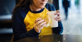 Kolorowa fotografia. Dziewczyna trzyma w ręce hamburgera. W drugiej trzyma kubek, z którego pije słodki, gazowany napój.